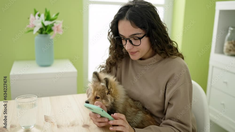 Young hispanic woman with dog sitting on table using smartphone at dinning room Young hispanic woman with dog sitting on table using smartphone at dinning room