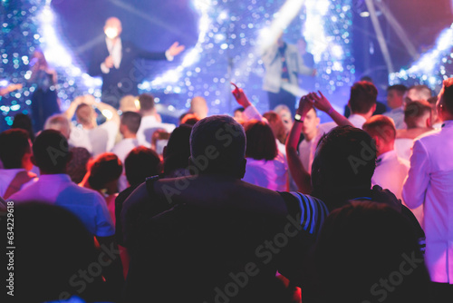 Crowded concert hall arena with scene stage lights with musicians band on a stage at the venue, rock show performance, with concert-goers attendees, audience on dance floor during concert festival