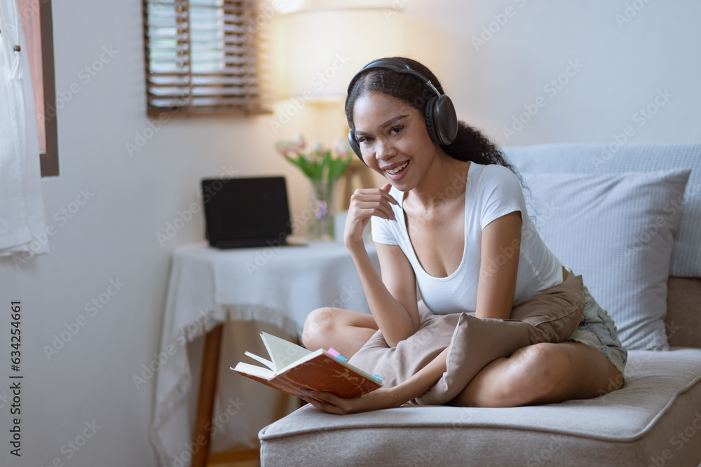 Young African woman holding a book with earphones sitting on sofa at home. Young African woman holding a book with earphones sitting on sofa at home.