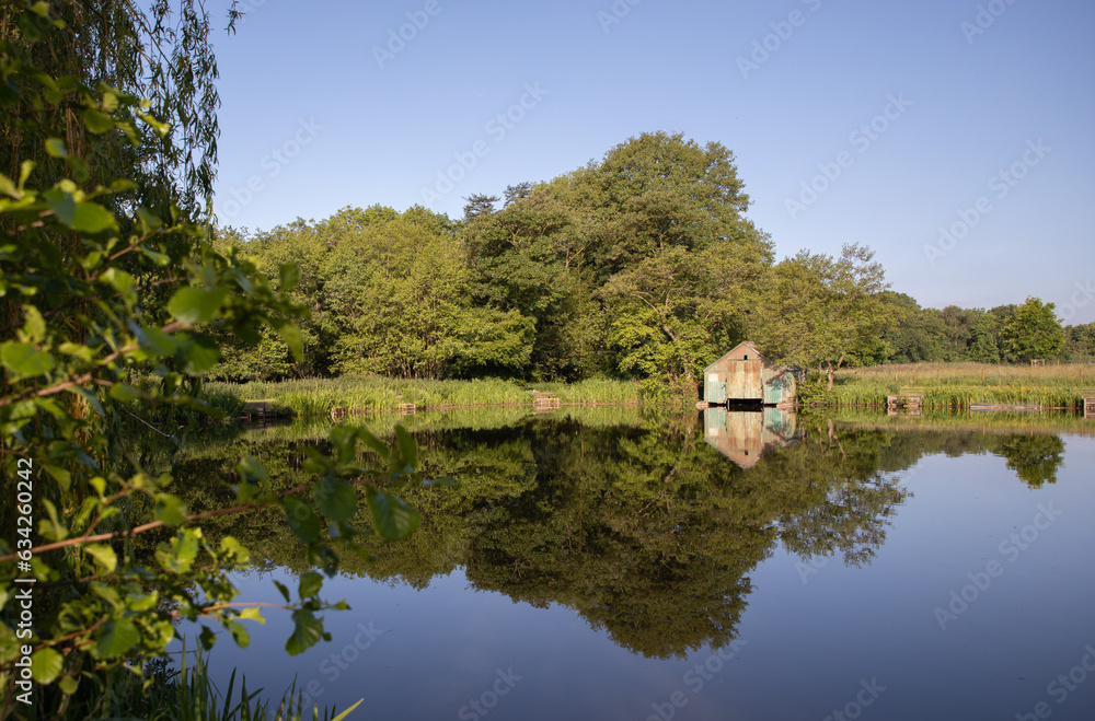 Taken on a sunny summer day with clear blue sky, it shows an old boat shed on a pool reflected in the water