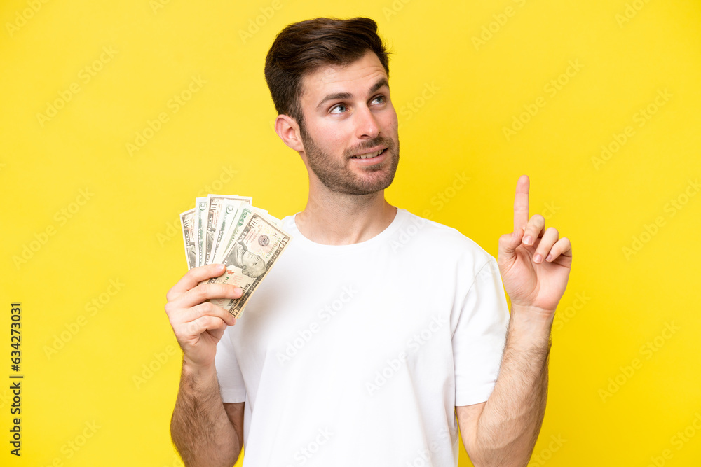 Young caucasian man taking a lot of money isolated on yellow background pointing up a great idea Young caucasian man taking a lot of money isolated on yellow background pointing up a great idea