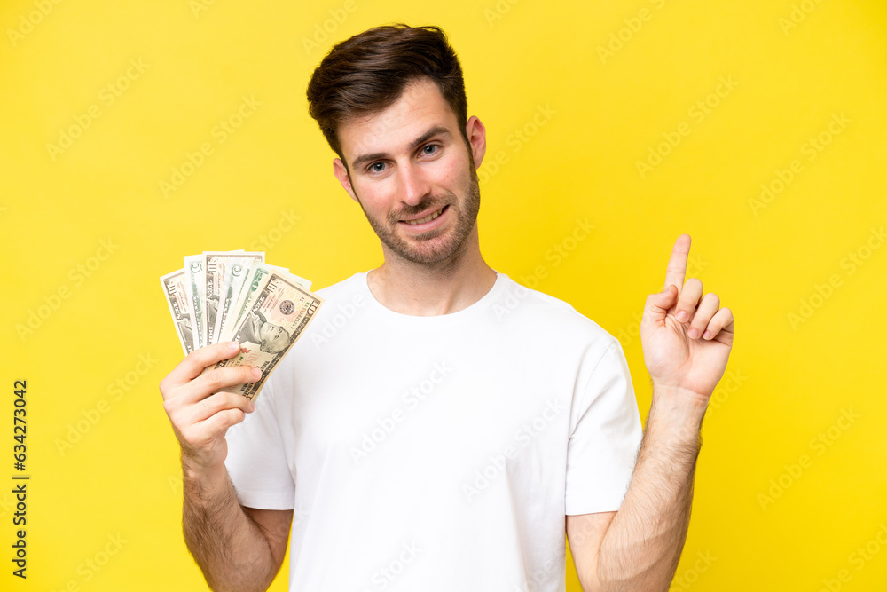 Young caucasian man taking a lot of money isolated on yellow background showing and lifting a finger in sign of the best Young caucasian man taking a lot of money isolated on yellow background showing and lifting a finger in sign of the best