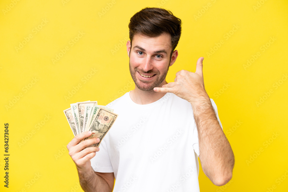 Young caucasian man taking a lot of money isolated on yellow background making phone gesture. Call me back sign Young caucasian man taking a lot of money isolated on yellow background making phone gesture. Call me back sign