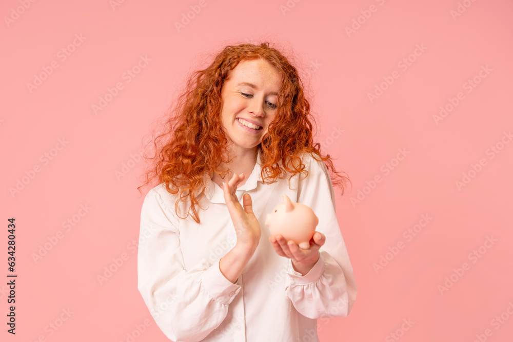 happy red hair woman in white shirt saving money for rainy day in the piggy bank, investment, saving money, currency, deposit. Indoor studio shot isolated with copy space on pink background. happy red hair woman in white shirt saving money for rainy day in the piggy bank, investment, saving money, currency, deposit. Indoor studio shot isolated with copy space on pink background.