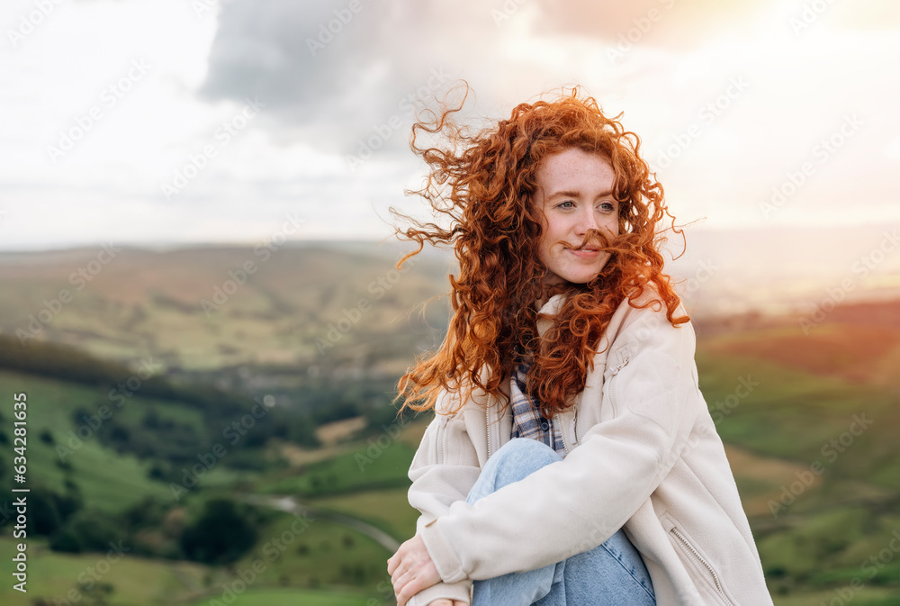 Happy smiling red hair Woman in jacket reaching the destination and sitting on the top of mountain at sunset. Travel Lifestyle concept The national park Peak District in England Happy smiling red hair Woman in jacket reaching the destination and sitting on the top of mountain at sunset. Travel Lifestyle concept The national park Peak District in England