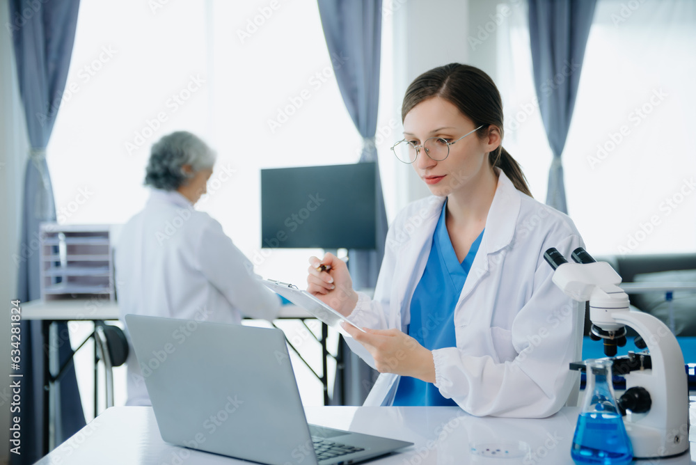 Confident young Caucasian female doctor in white medical uniform sit at desk working on computer. Smiling use laptop write in medical journal in clinic.. Confident young Caucasian female doctor in white medical uniform sit at desk working on computer. Smiling use laptop write in medical journal in clinic..