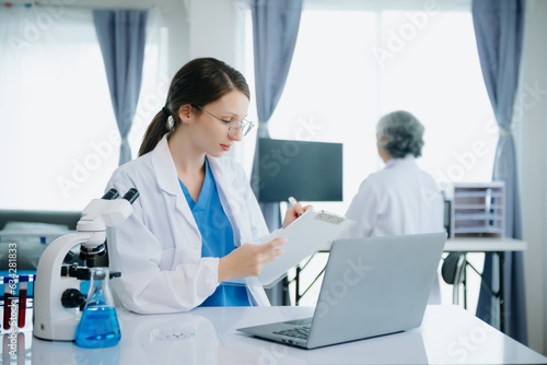 Fototapeta Naklejka Na Ścianę i Meble -  Confident young Caucasian female doctor in white medical uniform sit at desk working on computer. Smiling use laptop write in medical journal in clinic..