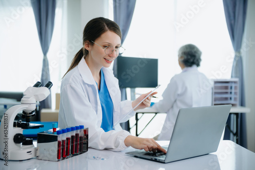 Confident young Caucasian female doctor in white medical uniform sit at desk working on computer. Smiling use laptop write in medical journal in clinic..