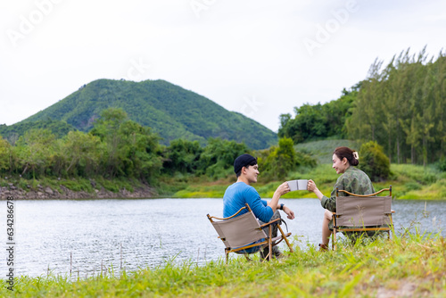 Asian man and woman friends relaxing by the lake drinking hot coffee together in the morning. People enjoy outdoor lifestyle travel nature hiking and camping in forest mountain on summer vacation.