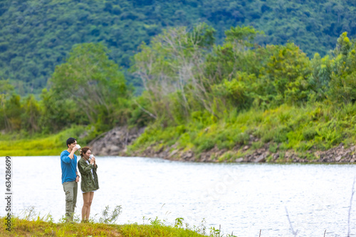 Asian man and woman friends relaxing by the lake drinking hot coffee together in the morning. People enjoy outdoor lifestyle travel nature hiking and camping in forest mountain on summer vacation.