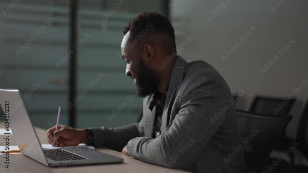 Side view portrait of smiling African-American businessman working typing keyboard on laptop computer and writing on note with pen sitting at table at office, with modern dark interior, slow motion. Side view portrait of smiling African-American businessman working typing keyboard on laptop computer and writing on note with pen sitting at table at office, with modern dark interior, slow motion.