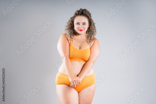 Curvy plus size model in orange underwear on gray background, happy overweight woman with fat buttocks, thighs, legs, hands and stomach, body positive concept