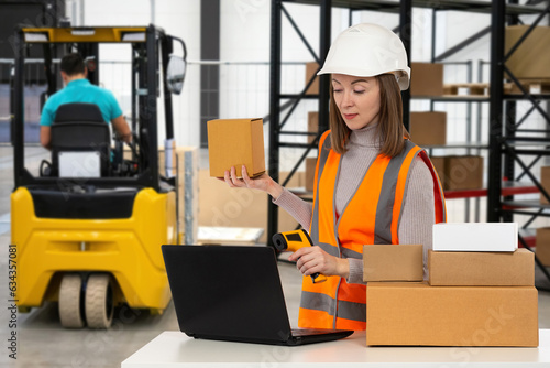 Storekeeper woman. Warehouse worker. Girl uses barcode scanner. Storekeeper is holding cardboard box. Woman works in warehouse. Forklift and shelving behind storekeeper. Data collection terminal