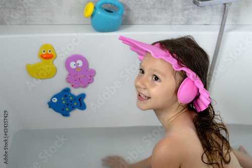 Fotografering A little smiling girl of European appearance with a funny bathing hat sits in the bathroom with toys