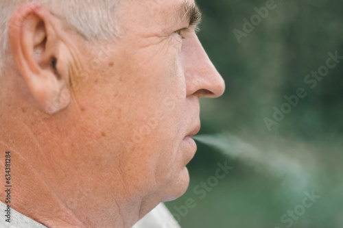 Senior man lighting up a cigarette outdoors against green nature background. Old thoughtful man smoking, looking aside. Close Up Face. Relaxed pensioner enjoy weekend rest outside. Unhealthy habits