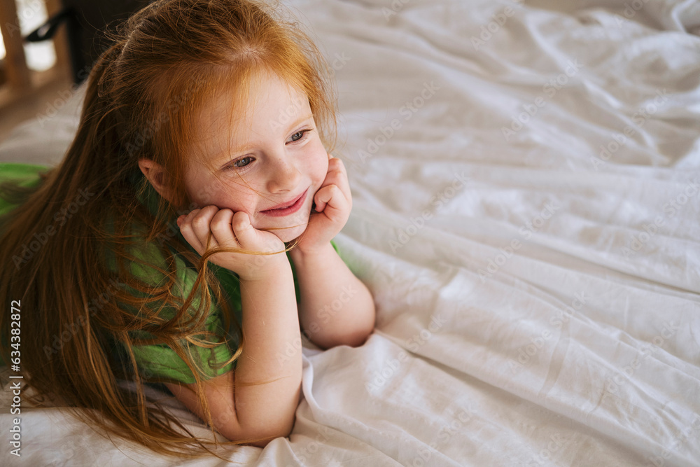 A red-haired little girl in a green T-shirt lies on a white bed at home and smiles. A red-haired little girl in a green T-shirt lies on a white bed at home and smiles.