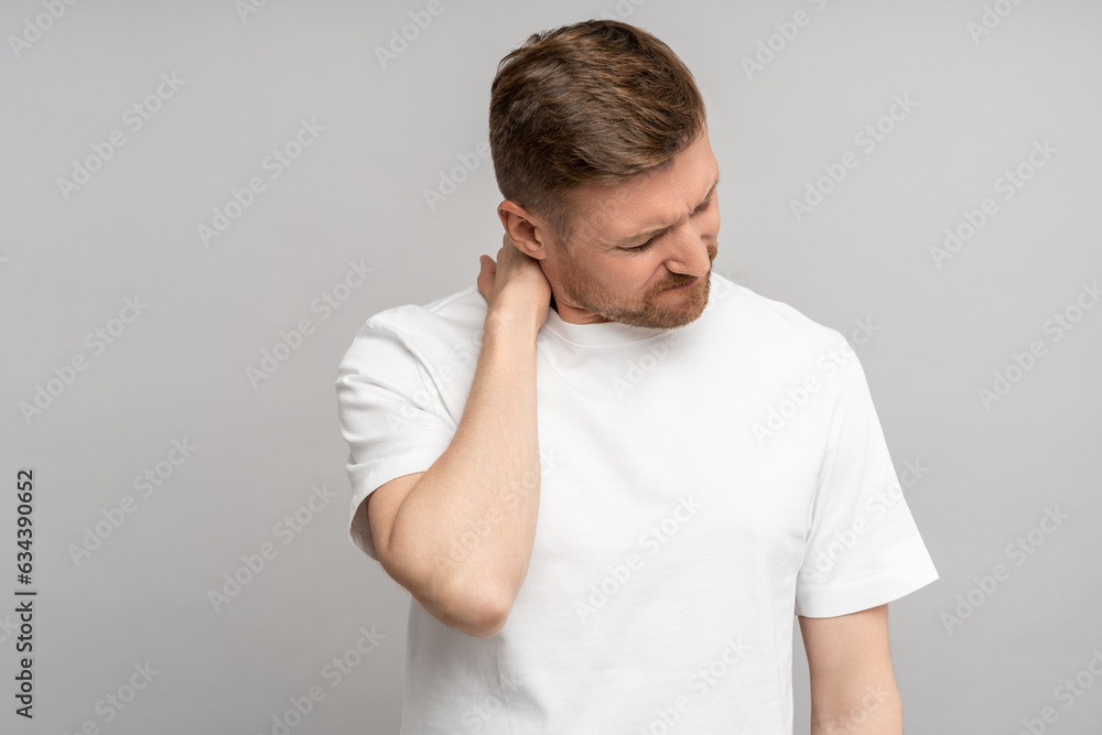 Unwell man frowns suffers from pain in neck isolated on grey studio wall. Inflammation of spinal discs hernia, sedentary lifestyle. Impact of overworking on male health. Physical problems in adulthood Unwell man frowns suffers from pain in neck isolated on grey studio wall. Inflammation of spinal discs hernia, sedentary lifestyle. Impact of overworking on male health. Physical problems in adulthood