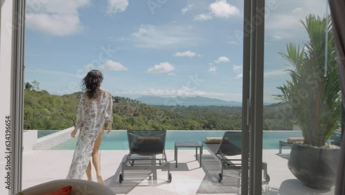 Stylish white summer house interior with open terrace and swimming pool on jungle background, woman in pareo walking out to lounge area, tropical holiday concept