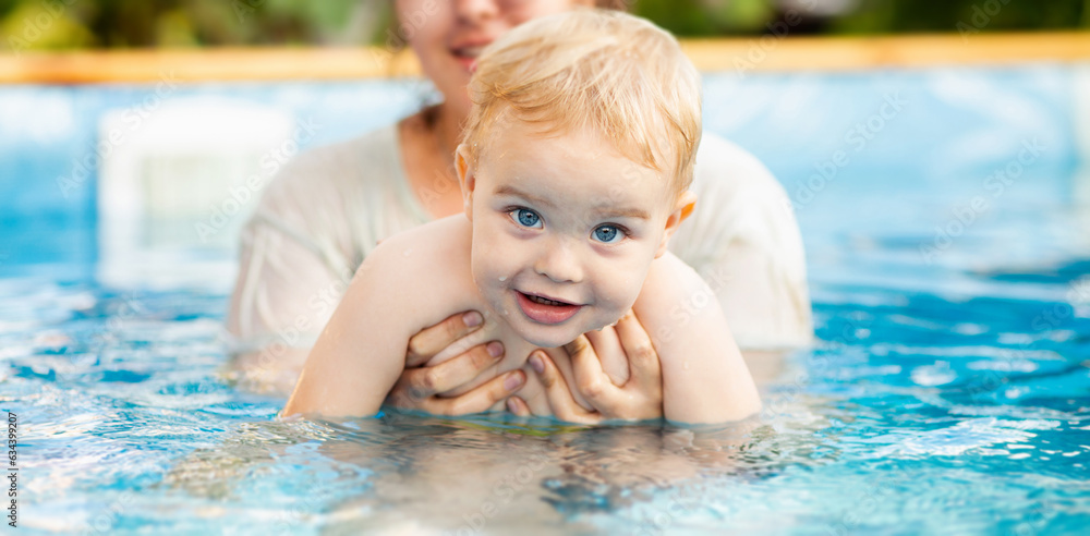 Portrait of small red-haired boy bathes in pool with hand support, baby swimming in water, summer leisure