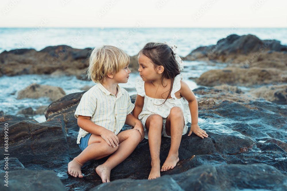 blond boy and a brunette girl, three years old, are sitting on stones on the seashore