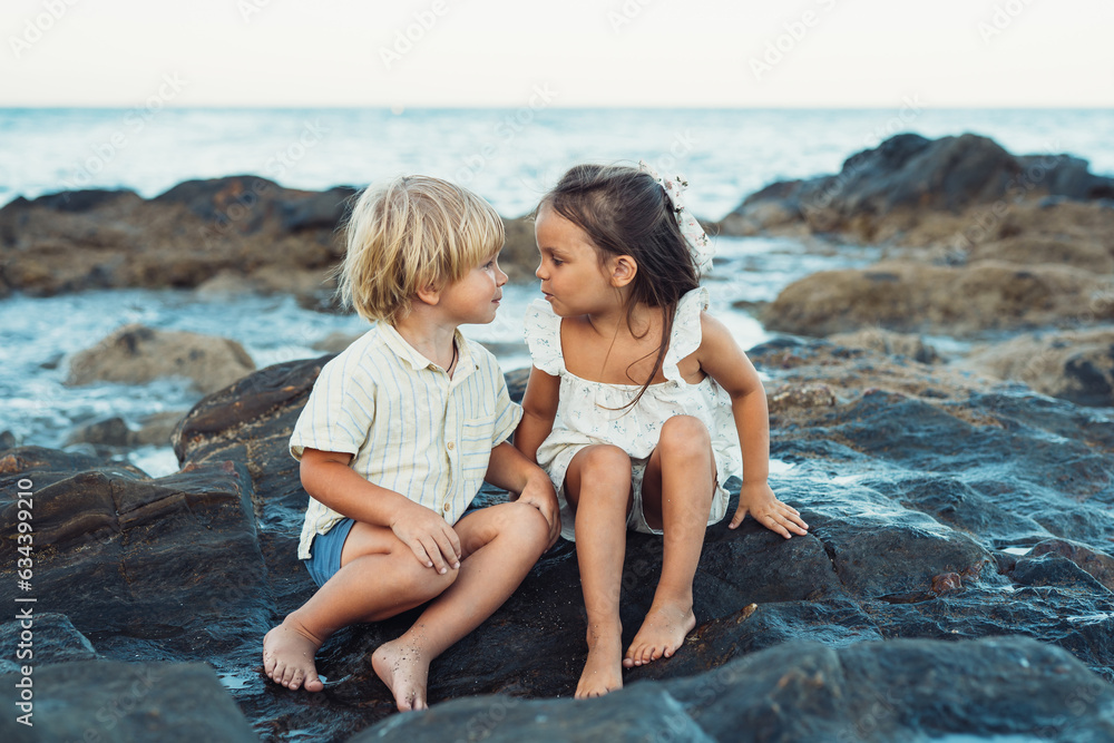 blond boy and a brunette girl, three years old, are sitting on stones on the seashore blond boy and a brunette girl, three years old, are sitting on stones on the seashore