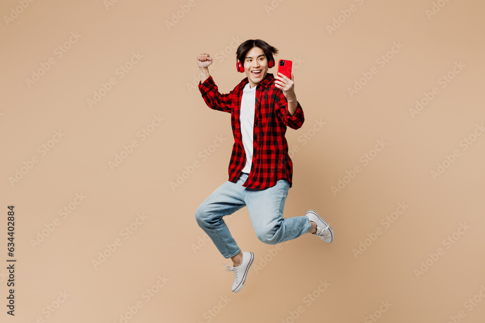 Full body side view young man of Asian ethnicity wear red shirt casual clothes jump high listen to music in headphones use mobile cell phone do winner gesture isolated on plain light beige background.