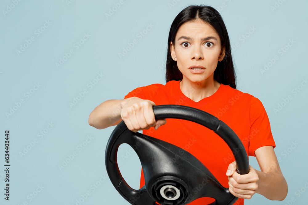 Young shocked astonished scared sad latin woman wear orange red t-shirt casual clothes hold steering wheel driving car look camera isolated on plain pastel blue background studio. Lifestyle concept. Young shocked astonished scared sad latin woman wear orange red t-shirt casual clothes hold steering wheel driving car look camera isolated on plain pastel blue background studio. Lifestyle concept.