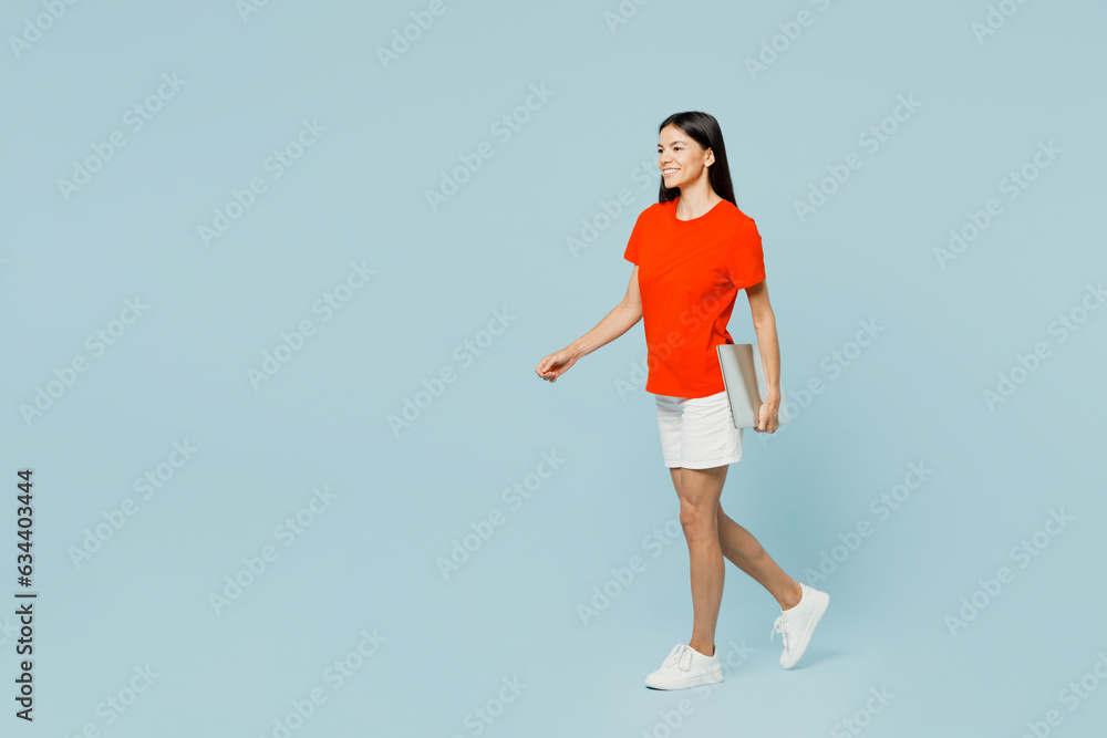 Full body side view young latin woman wear orange red t-shirt casual clothes hold closed laptop pc computer walking going isolated on plain pastel light blue cyan background studio. Lifestyle concept. Full body side view young latin woman wear orange red t-shirt casual clothes hold closed laptop pc computer walking going isolated on plain pastel light blue cyan background studio. Lifestyle concept.