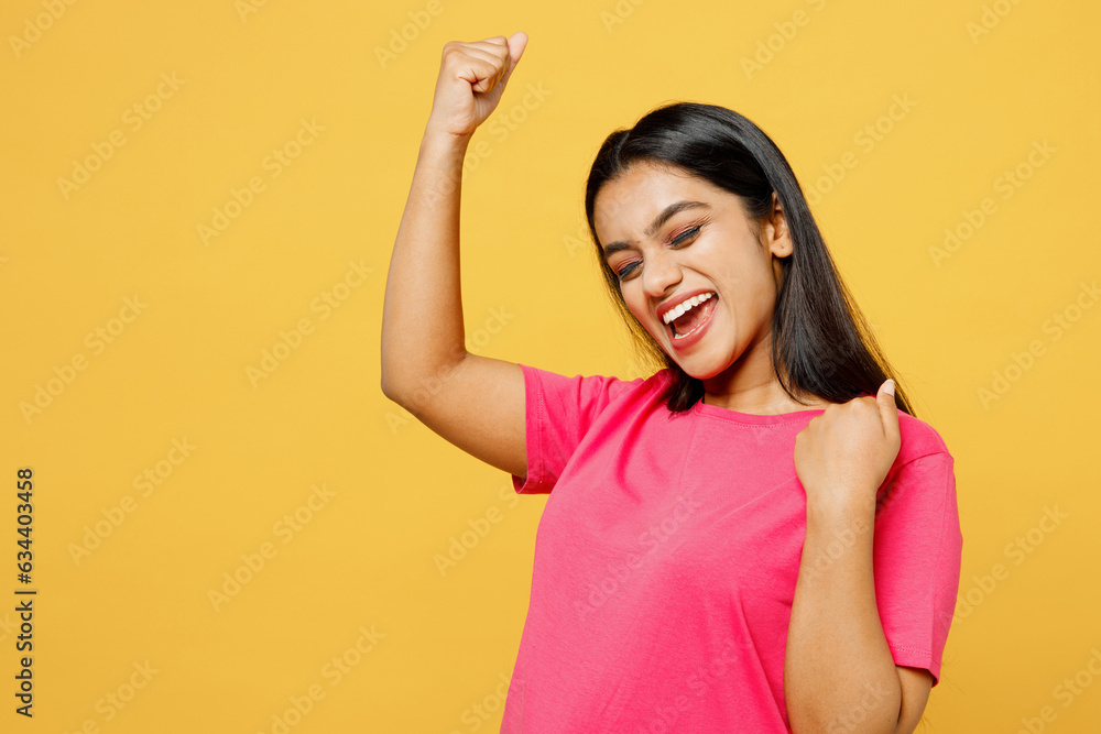 Young overjoyed fun happy Indian woman wear pink t-shirt casual clothes doing winner gesture celebrate clenching fists say yes isolated on plain yellow background studio portrait. Lifestyle concept Young overjoyed fun happy Indian woman wear pink t-shirt casual clothes doing winner gesture celebrate clenching fists say yes isolated on plain yellow background studio portrait. Lifestyle concept