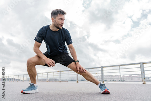 A trainer in fitness clothes is a T-shirt, running sports shoes and a smart watch for sports.    A strong man. A confident male athlete training does warm-up exercises. © muse studio