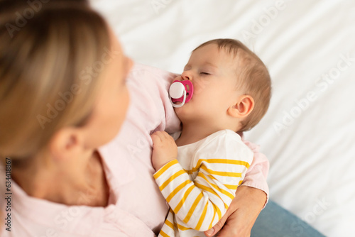Caring mother holding baby girl sleeping with pacifier, comforting kid and singing lullaby, sitting on bed, above view