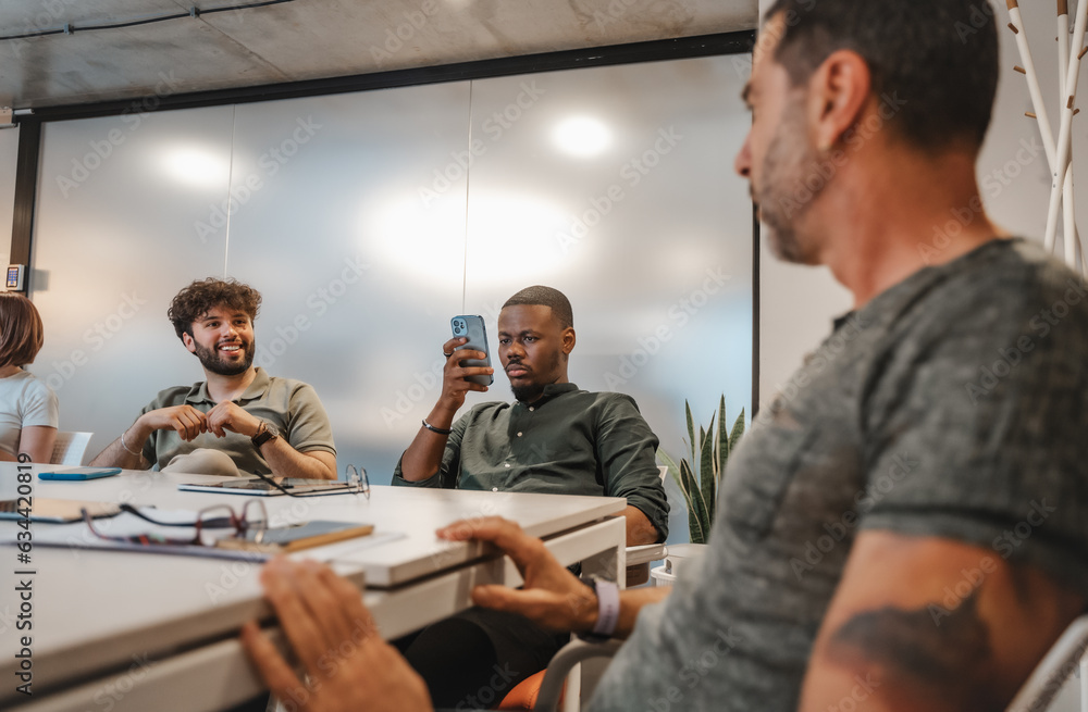 Fototapeta premium Good looking black male person scrolling on his phone while his colleagues are having a conversation