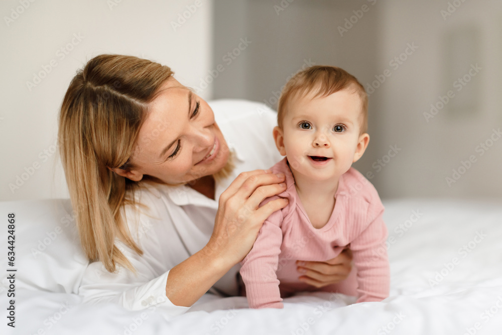 Loving mother talking and playing to her infant child girl, kid crawling and smiling at camera on bed in bedroom
