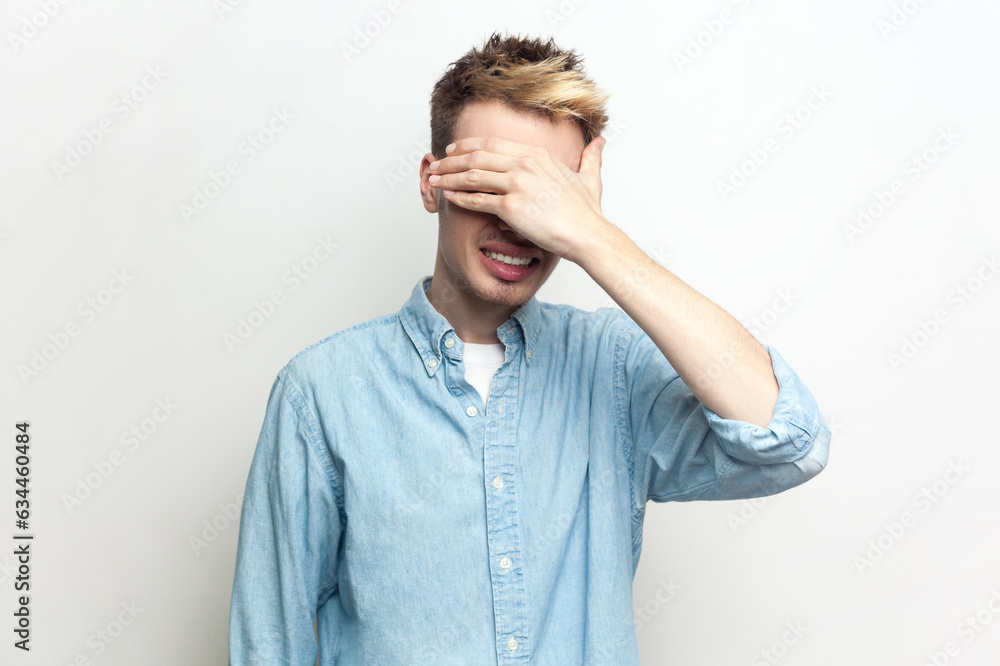 Portrait of shocked scared man wearing denim shirt standing covering his eyes with palm, doesn't want to see something frighten. Indoor studio shot isolated on gray background. Portrait of shocked scared man wearing denim shirt standing covering his eyes with palm, doesn't want to see something frighten. Indoor studio shot isolated on gray background.