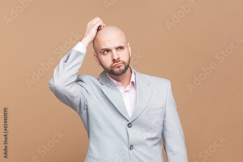 Portrait of indignant puzzled bald bearded man rubbs head, tries to remember necessary information, looks in displeasure, wearing gray jacket. Indoor studio shot isolated on brown background.