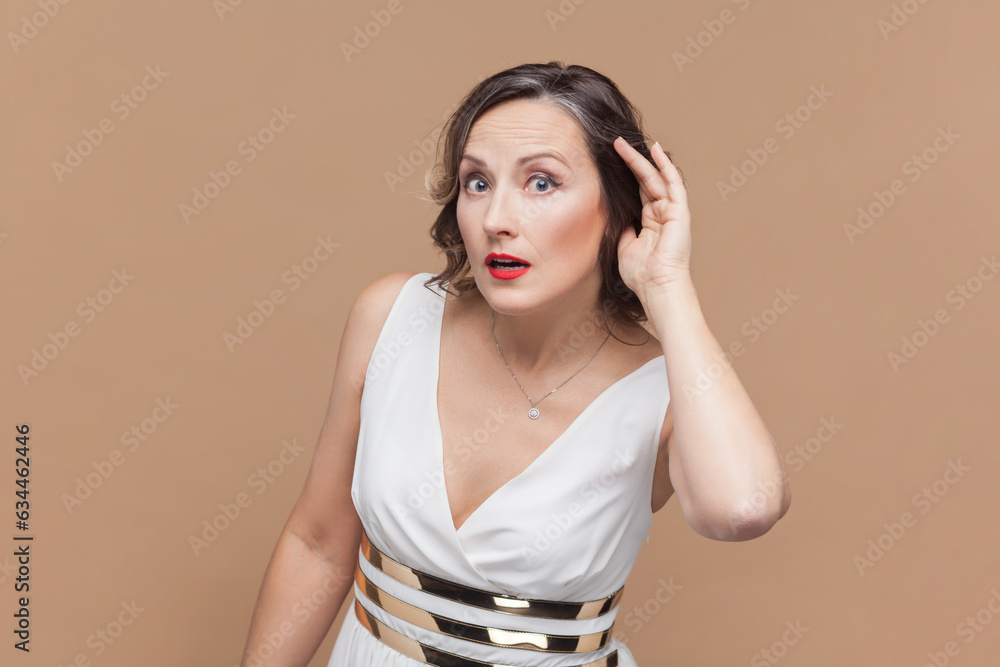Can't hear you. Attentive middle aged woman holding hand near ear trying to listen quiet conversation, overhearing gossip, having hearing problems Indoor studio shot isolated on light brown background