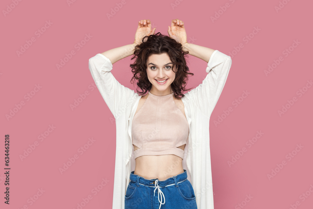 Portrait of funny childish woman with curly hair wearing casual style outfit having fun, making bunny ears with palms, looking smiling at camera. Indoor studio shot isolated on pink background. Portrait of funny childish woman with curly hair wearing casual style outfit having fun, making bunny ears with palms, looking smiling at camera. Indoor studio shot isolated on pink background.