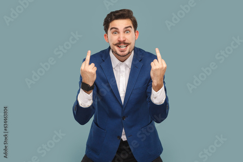 Rude impolite handsome man with mustache standing showing middle fingers, looking at camera with clenched teeth, wearing white shirt and jacket. Indoor studio shot isolated on light blue background.
