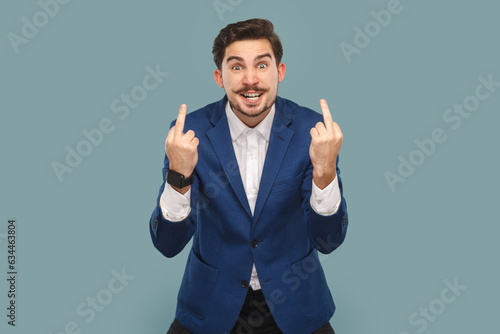 Rude impolite handsome man with mustache standing showing middle fingers, looking at camera with clenched teeth, wearing white shirt and jacket. Indoor studio shot isolated on light blue background. photo