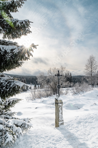 Cross and the winter snowy landscape at Filipova Hut, Sumava national park, sun shining behind the trees