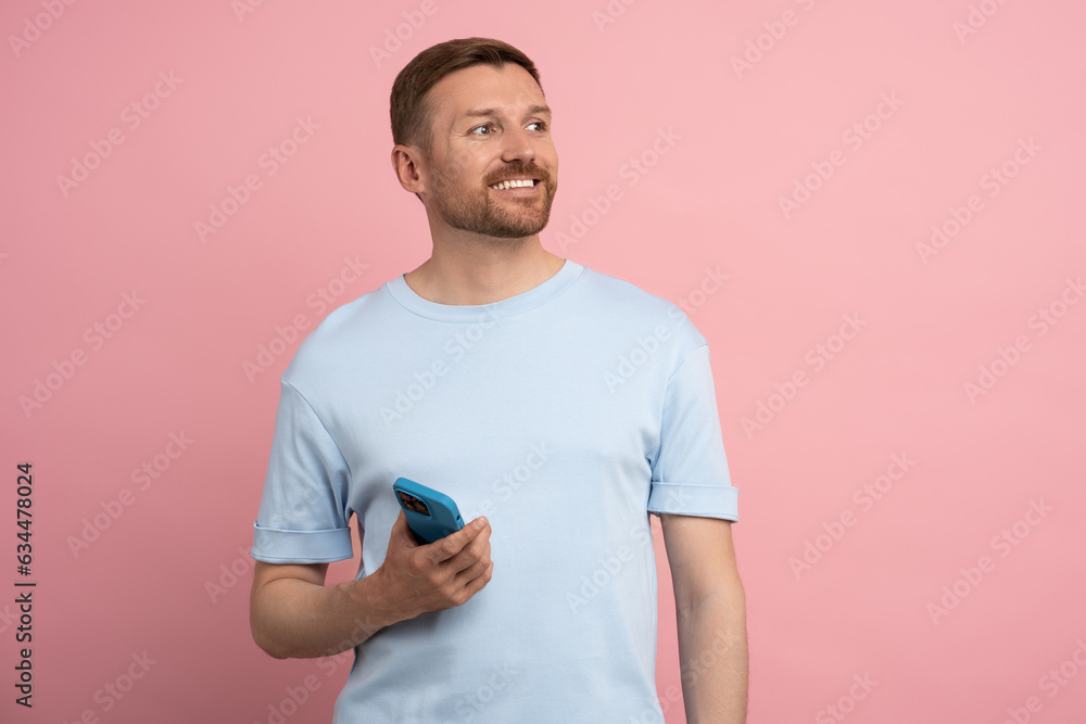 Smiling happy man looks away on empty wall space holding mobile phone against wall in studio room. Interested guy rejoices having good mood from message offer by cellphone isolated pink background. Smiling happy man looks away on empty wall space holding mobile phone against wall in studio room. Interested guy rejoices having good mood from message offer by cellphone isolated pink background.