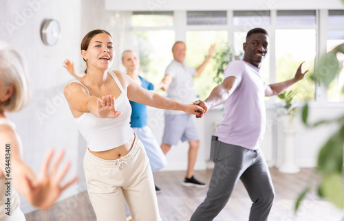 In fitness studio, young girl teacher paired with african man dancing Latin American dance. Clients of different ages are engaged in fitness in background