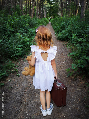 Beautiful little girl in a white dress with a toy and a suitcase alone in the forest. Concept: the child left home due to problems in the family, the baby was lost. photo