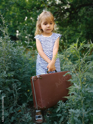 Beautiful little girl in a white dress with a toy and a suitcase alone in the forest. Concept: the child left home due to problems in the family, the baby was lost. photo