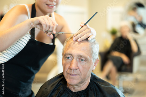 Cropped unrecognizable young woman hairdresser performs haircut for elderly man using scissors. Blurry senior woman waiting in background