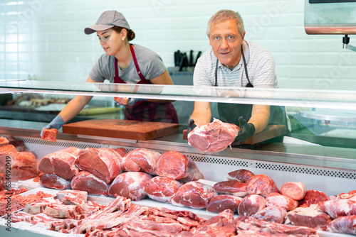 Adult man and young woman sellers in uniform display raw meat beef in butcher shop..