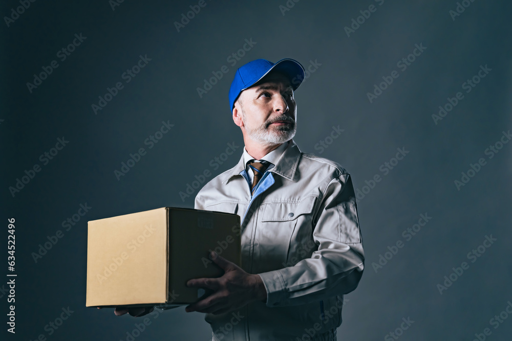 Portrait of an elderly Caucasian delivery staff in work clothes holding a cardboard box luggage. Portrait of an elderly Caucasian delivery staff in work clothes holding a cardboard box luggage.