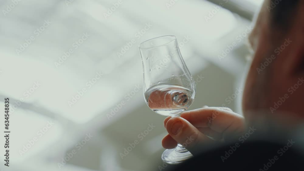 Male analyst holding a glass with distilled gin while checking it, industrial process in a gin distillery production Male analyst holding a glass with distilled gin while checking it, industrial process in a gin distillery production