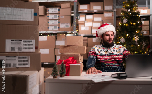 Online store inventory manager Santa Claus using laptop computer, working in storeroom on christmas sale, Young confident small business owner preparing order for client.