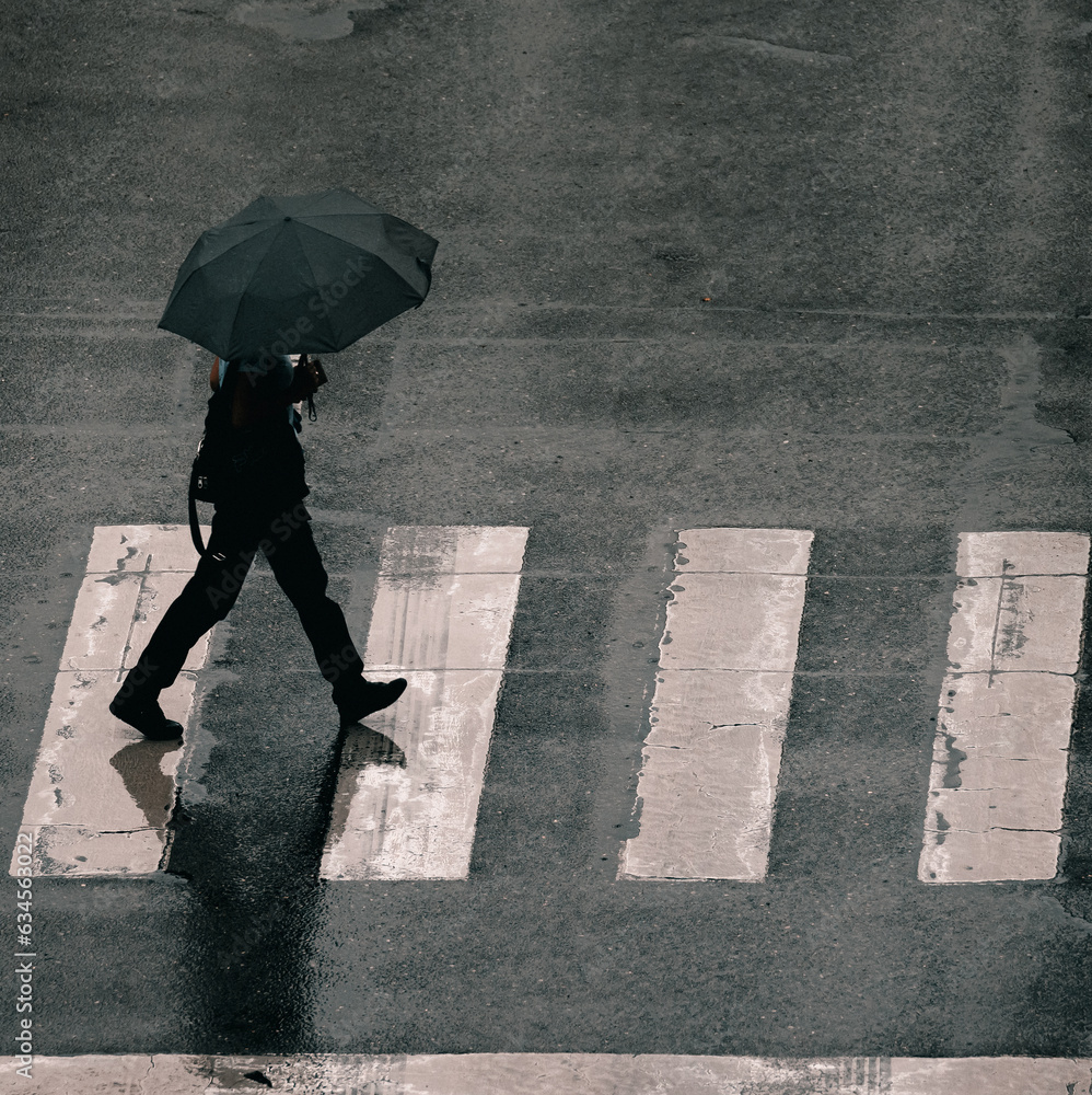 silhouette of a person walking with umbrella at a crossing - obrazy, fototapety, plakaty 